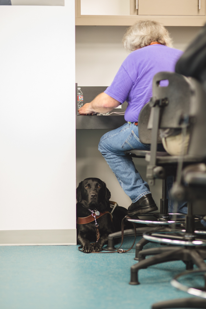 A man with long gray hair and a purple shirt sits facing away at one of the high top work surfaces in the Innovation Lab. His glossy black guide dog is on the floor at his feet, staring directly into the camera.