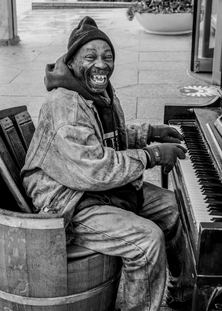 Photo 5: The same man breaks into a toothy grin. The shot is farther away and reveals the piano he sits at, his gloved finger pressing into ivory keys. Click the image to hear the corresponding audio file.