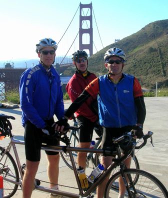 Chris, his tandem partner and another companion are standing before a view of the Golden Gate Bridge in their biking gear, with their bikes