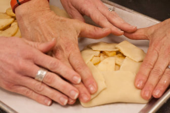 Two pairs of hands fold pastry dough around an apple filling.
