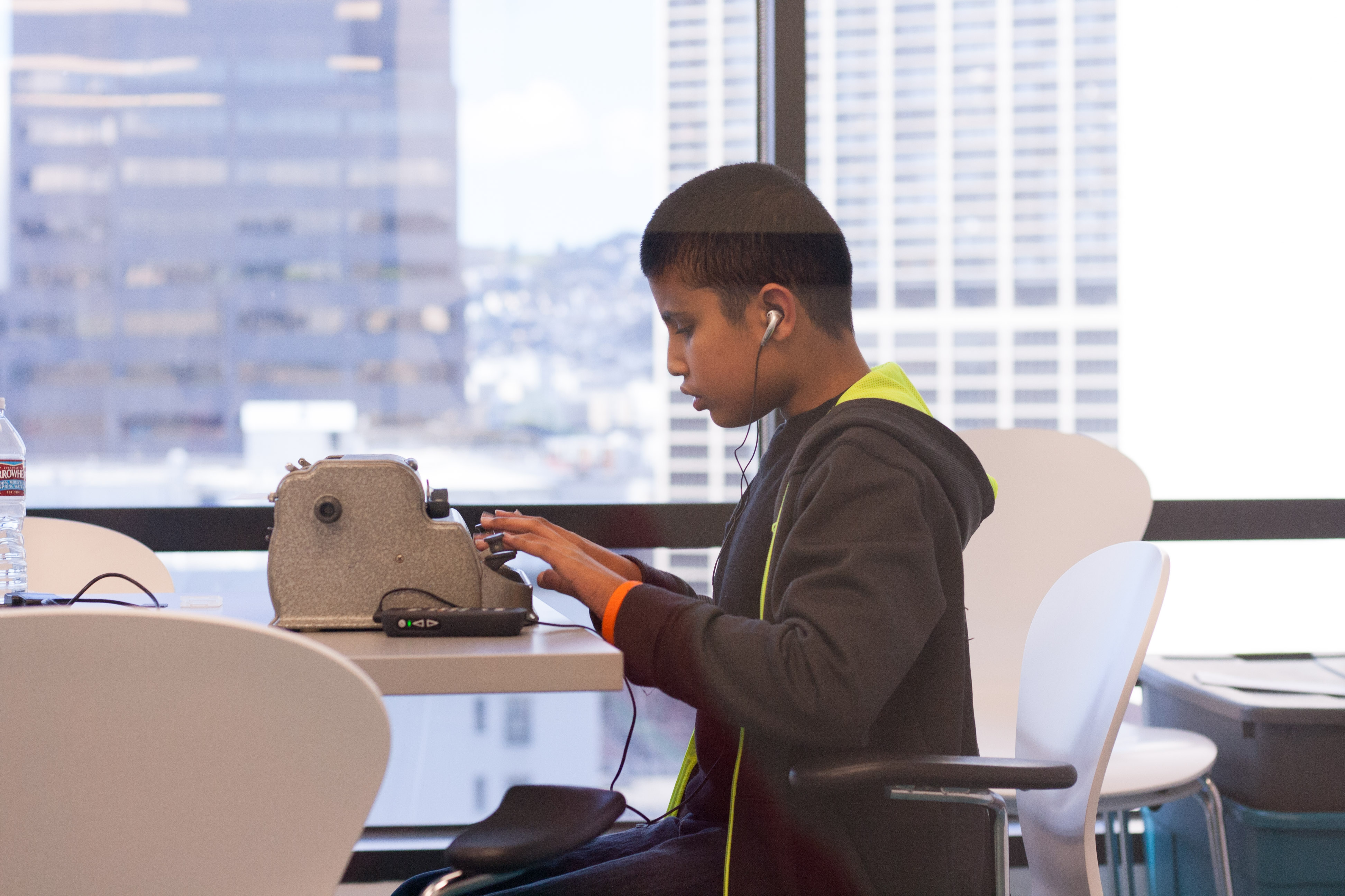 Competitor Rasheed sits typing at his brailler with headphones in. He is silhouetted against large windows that show the buildings of San Francisco in the background.