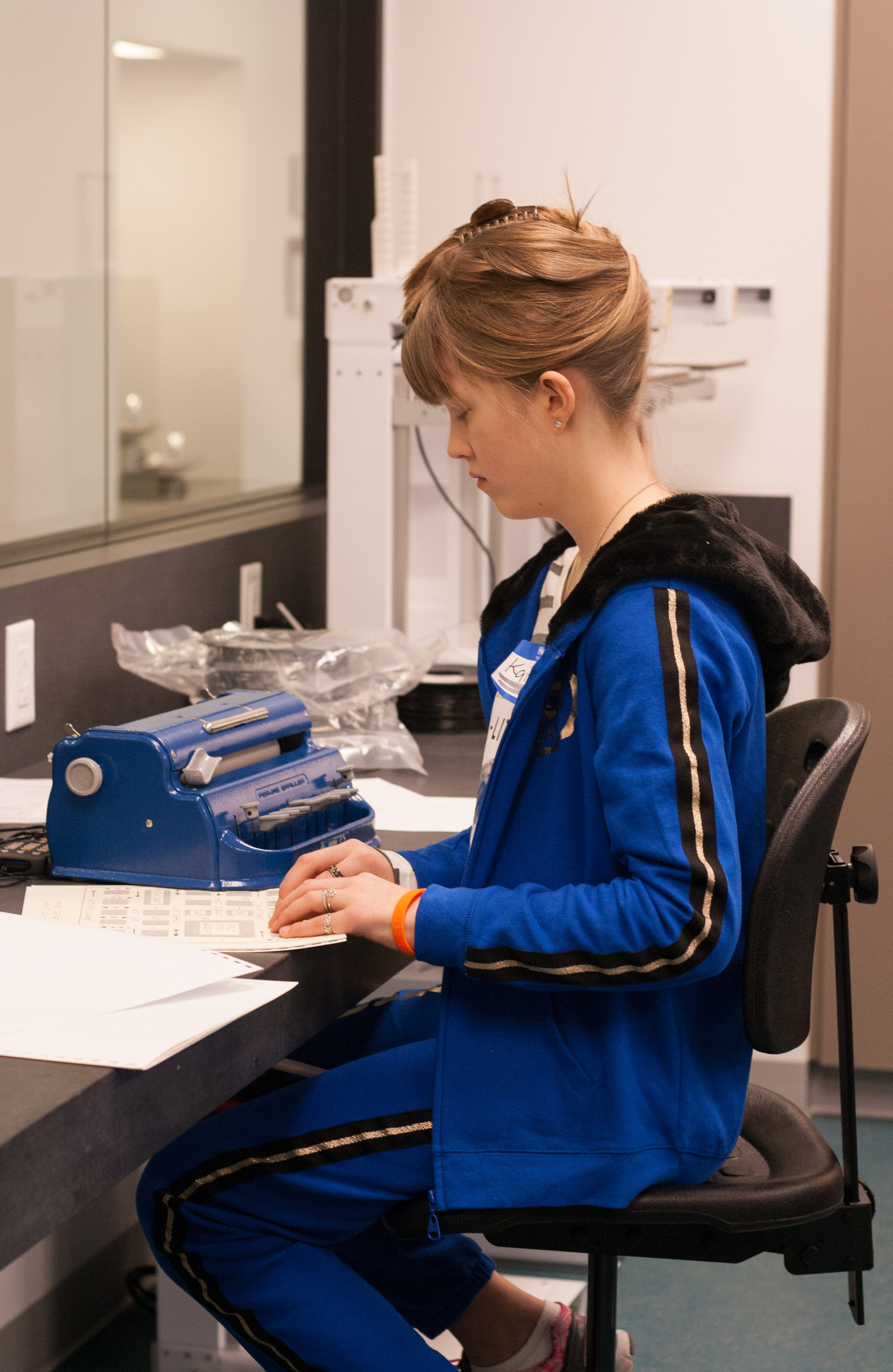 Junior Varsity competitor Kaitlyn reads a braille chart. She is wearing a royal blue tracksuit that nicely matches her royal blue brailler.