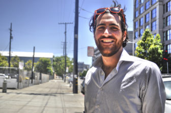 A man standing before a long sidewalk smiles with sunglasses perched on his head.