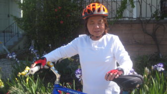 A woman dressed in white with a orange helmet stands with her bike steadied in front of her. 