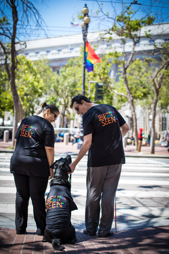LightHouse staff member Esmeralda Soto and Ethan Meigs cross the street wearing LightHouse's 'Be Seen' Pride Tshirts. The shirts say 'be seen' in orange uppercase letters and 'SF Pride 2017', with a braille 'L' and 'H' in rainbow. 