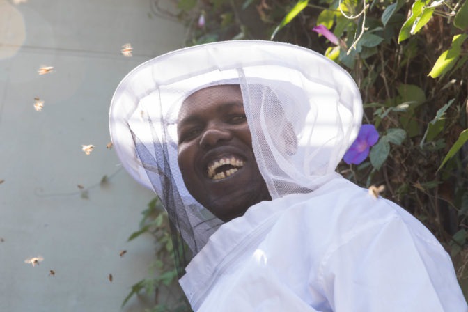 Ojok smiles with his white bee hat and net catching the light, while bees fly around him.