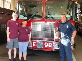 Chris Lawyer and Jess Marenoff, wearing their maroon EHC t-shirts, pose with firefighter Alexander Hermann in front of the grill of a giant red firetruck. 