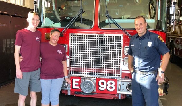 Chris Lawyer and Jess Marenoff, wearing their maroon EHC t-shirts, pose with firefighter Alexander Hermann in front of the grill of a giant red firetruck. Chris and Jess gave Hermann EHC t-shirts for the heroic crew.