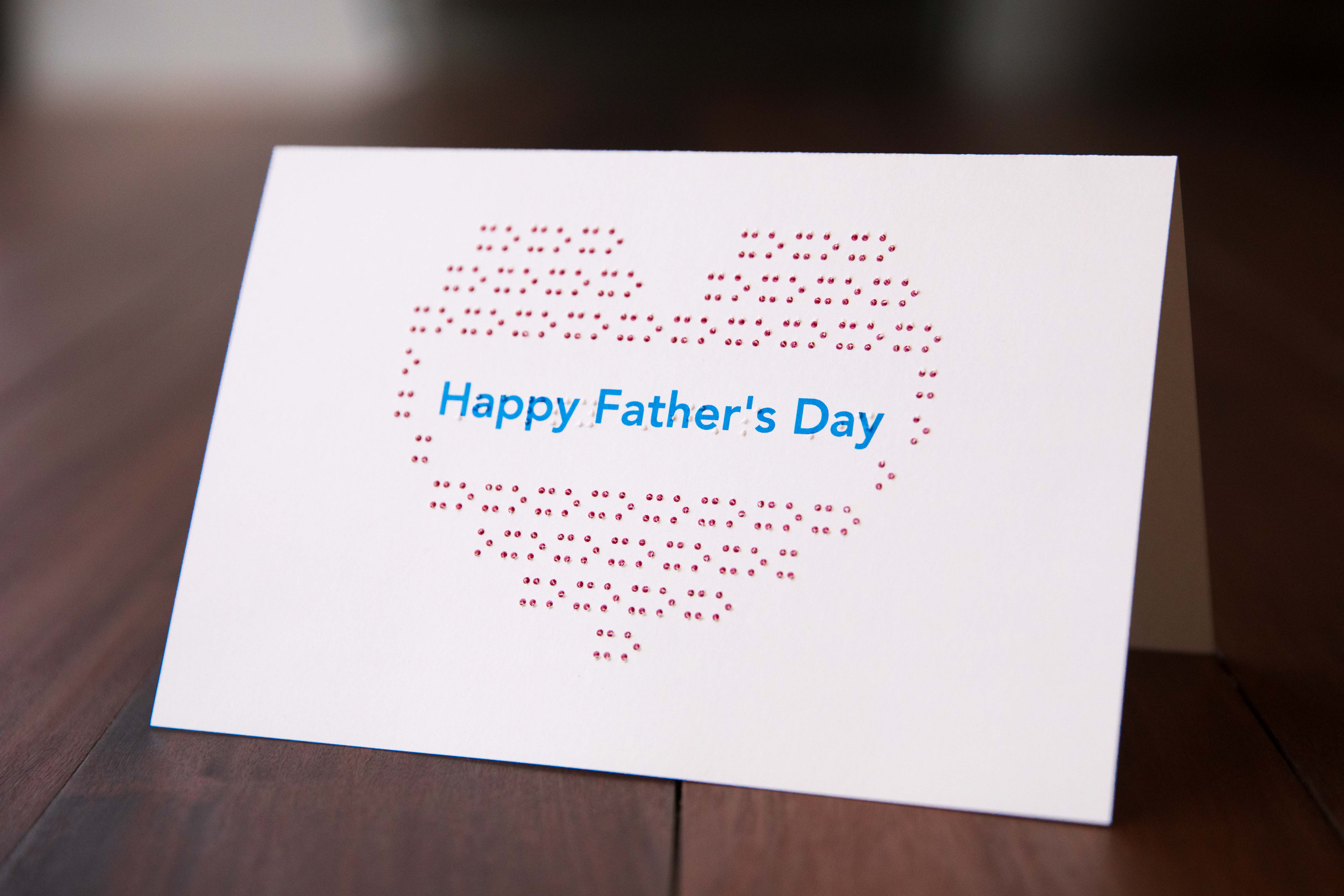The words 'Happy Father’s Day’ in blue text and braille, in the middle of a red ink and tactile heart, made up of braille x’s and o’s.