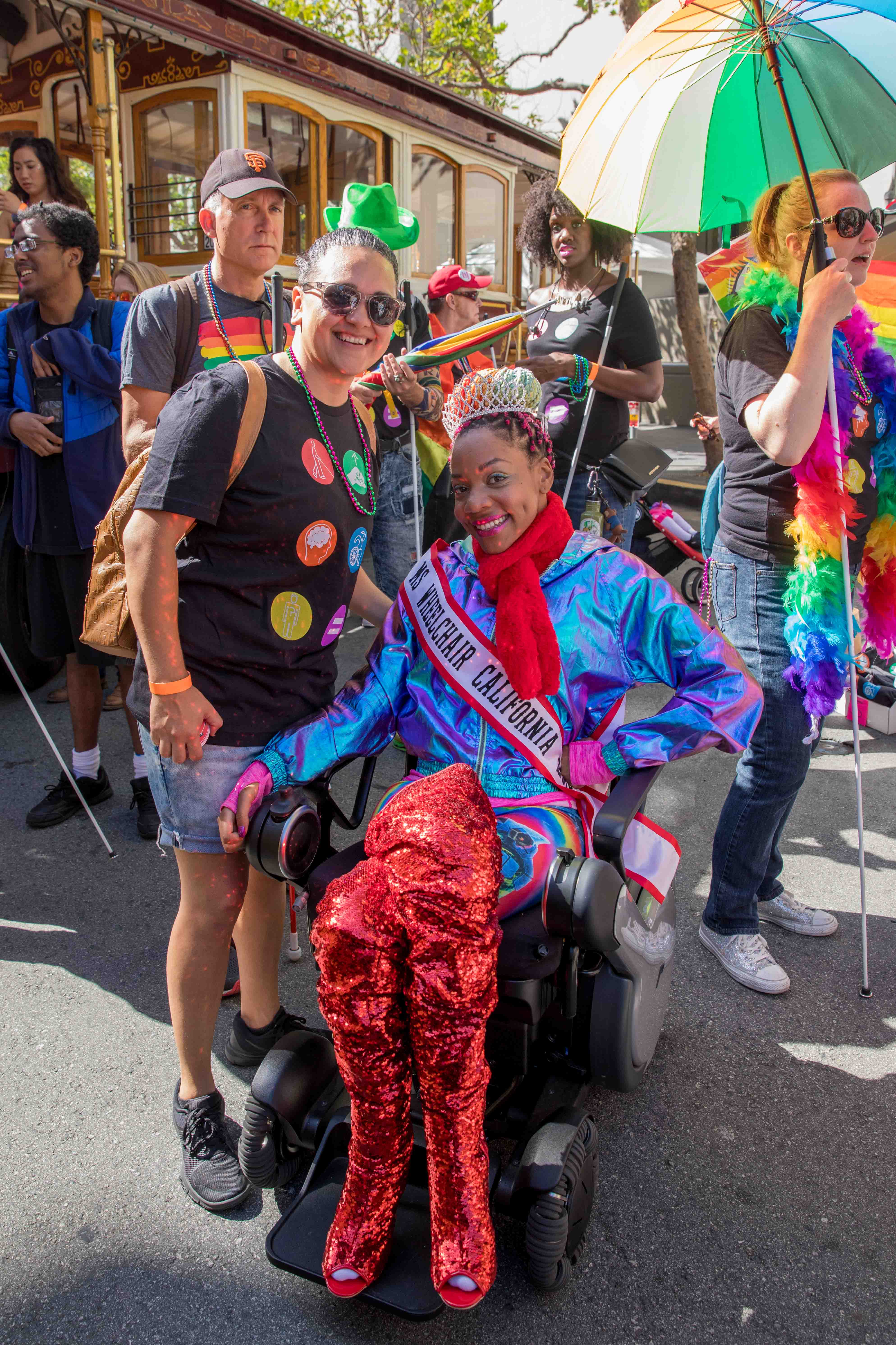 Two pride participants, one standing wearing the LightHouse shirt and wearing 'Ms. Wheelchair California' sash, prepare to march in the parade.
