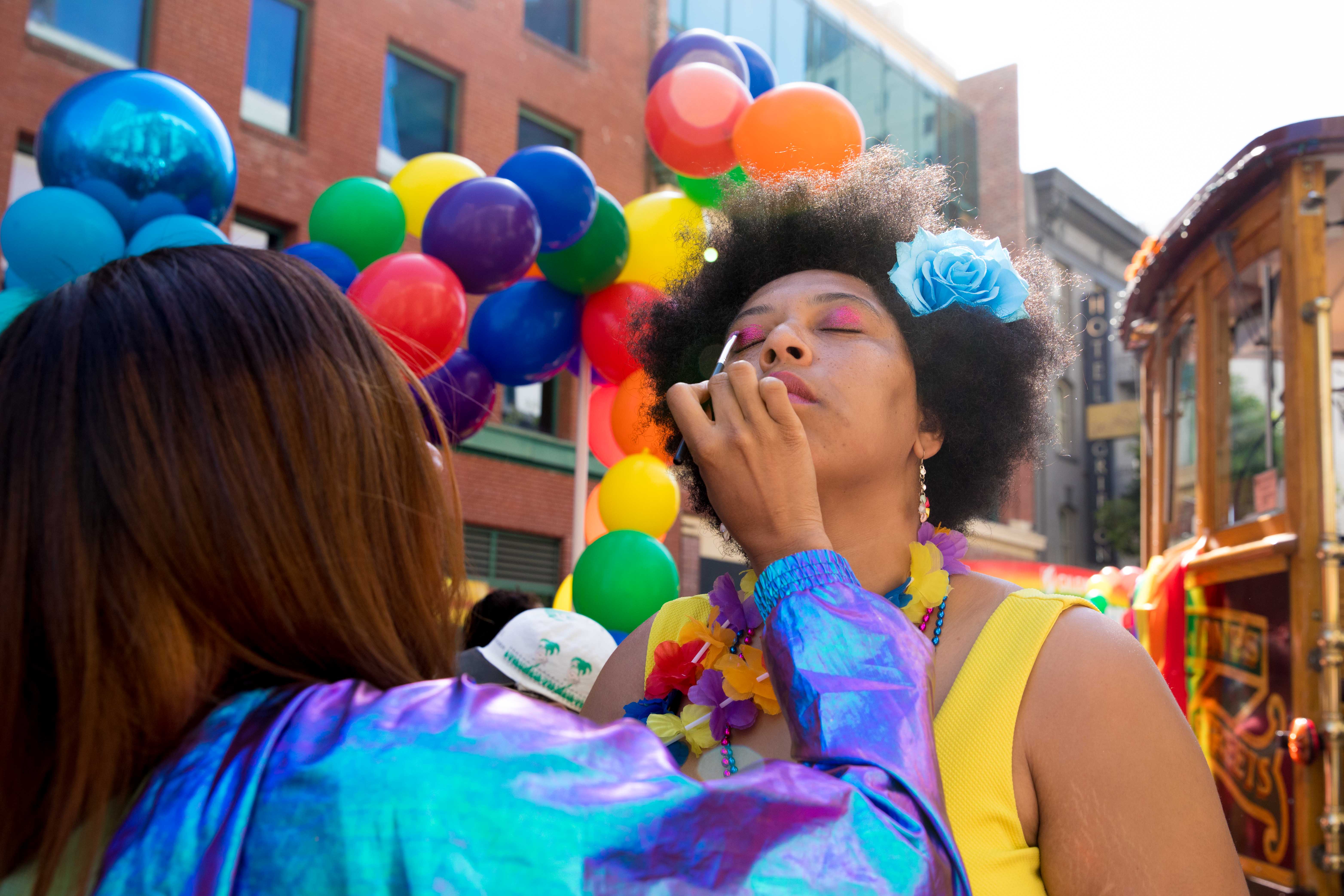A woman applies eyeshadow to a LightHouse contingent member with rainbow balloons in the background.