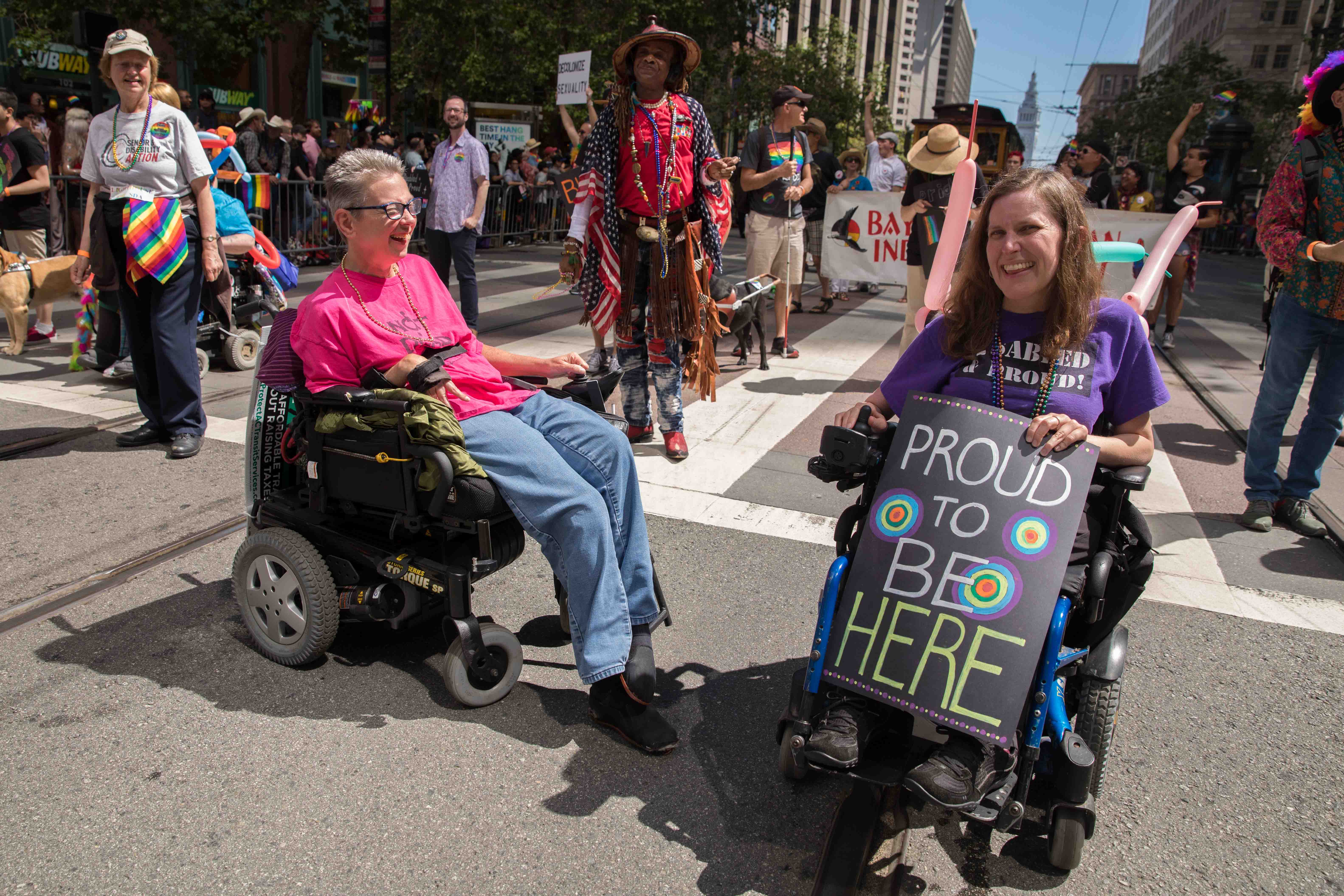 Two Pride participants in wheelchairs laugh while marching down market street with the contingent. One holds a sign that reads "Proud to be here."