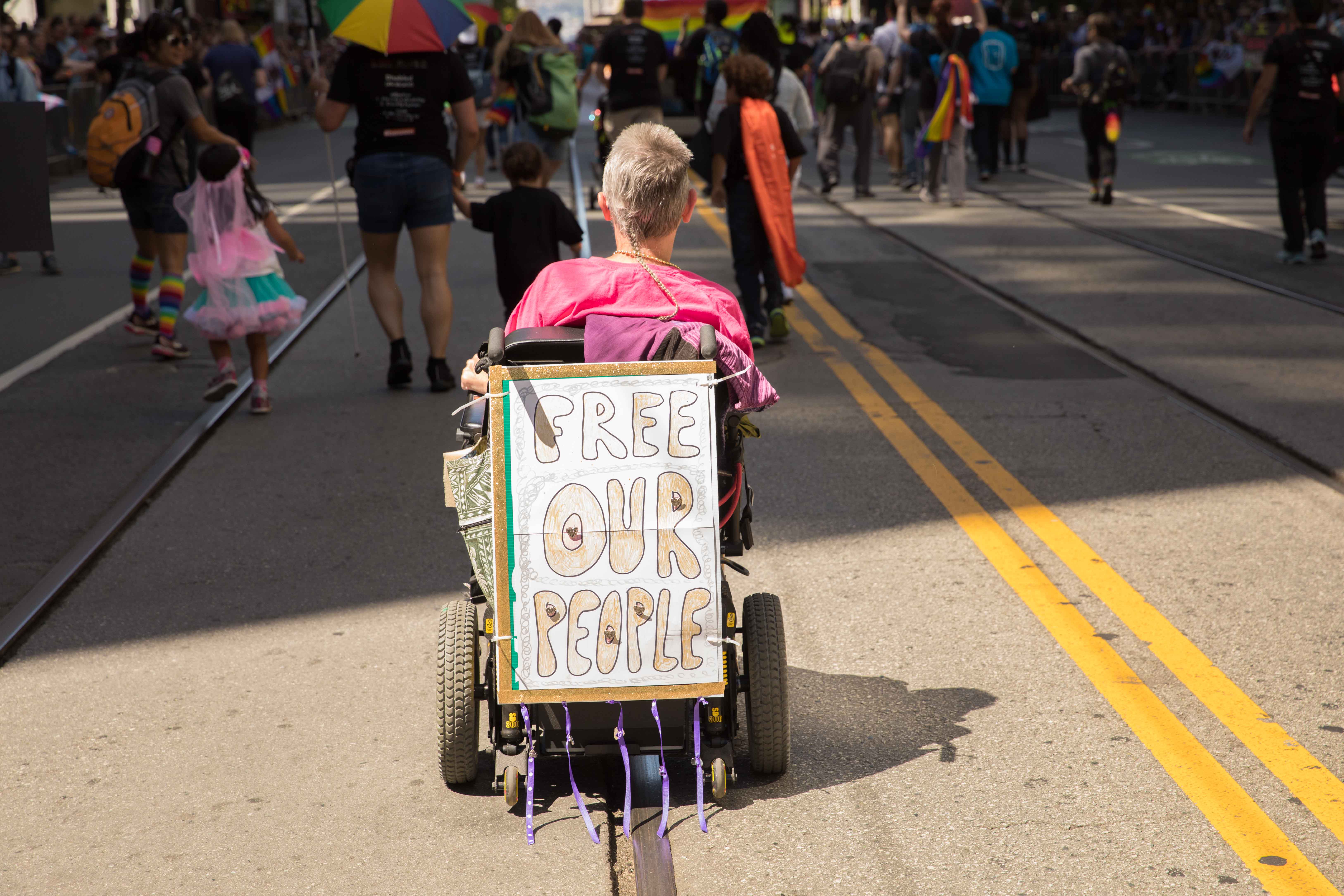 A contingent member marches with a sign attached to their wheelchair that reads "Free our people." 