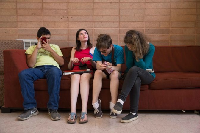 Four teen campers chat and look at their phones while sitting on a couch.