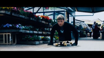 A young man rolls with his torso on a skateboard and arms pushing him past flowers in a market.