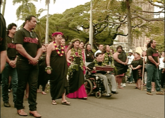A group of people walk in a march, and one joins in a wheelchair. The people wear leis and flowers, and men wearing shirts that say "Security" surround at the bounds.