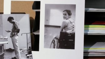 Two photographs sit upright beside a shelf of papers. At left, a man stands looking at his phone. In the photo at right, a woman sitting in a wheelchair smiles back at the camera.