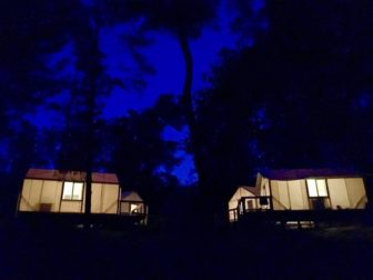 Two bungalows sit illuminated next to each other in the evening woods.