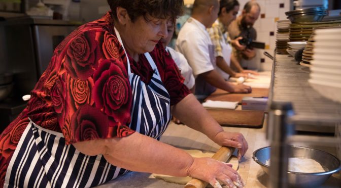 Penny Melville-Brown, wearing a shirt covered in red roses and a striped apron, rolls out a piece of dough in a kitchen surrounded by other bakers.