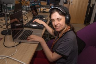 Audio Academy student Jenna smiles, seated, with one hand on her laptop and the other on her DJ equipment.