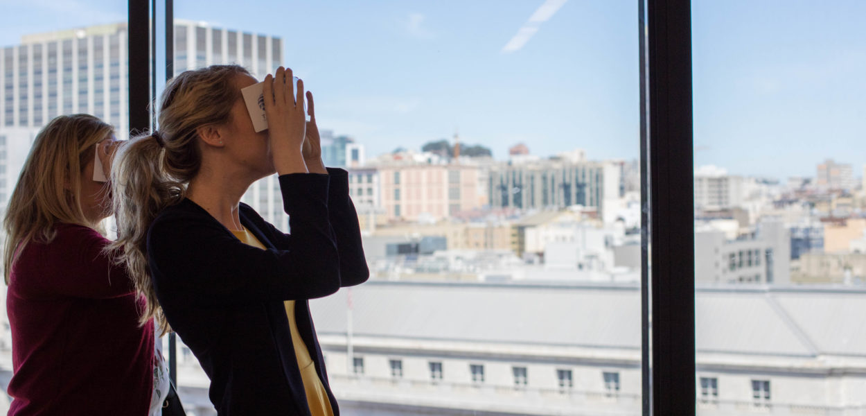 A woman tries on a Virtual Reality headset with the San Francisco skyline visible through the windows behind her.