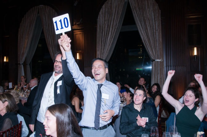 An auction participant raises his number triumphantly, donning a wide grin, amidst a crowd of cheering Gala attendees.
