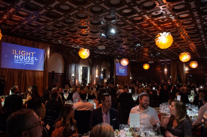 A medium shot of the Julia Morgan Ballroom during the Gala, with full tables of people, and illuminated screens with a graphic that says "LightHouse for the Blind and Visually Impaired".