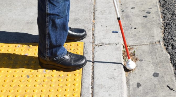 A shot of the shoes and tip of the cane of a person standing at a crosswalk, using a white cane.