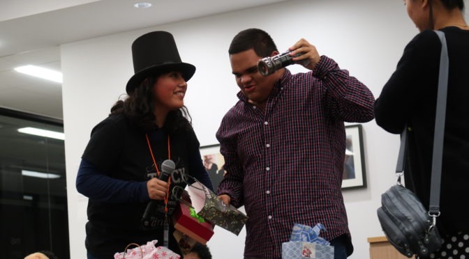A person wearing a top hat and holding a microphone smiles as another holds an opened gift up to their ear at the 2018 LightHouse Youth White Elephant Gift Exchange.