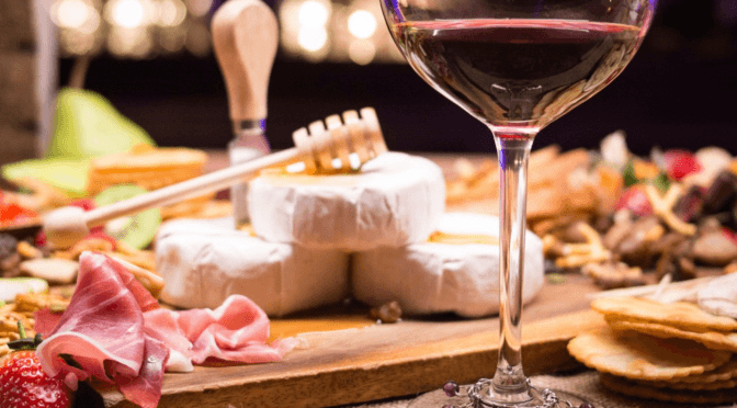 A glass of wine on a board of cheese and meat and fruit