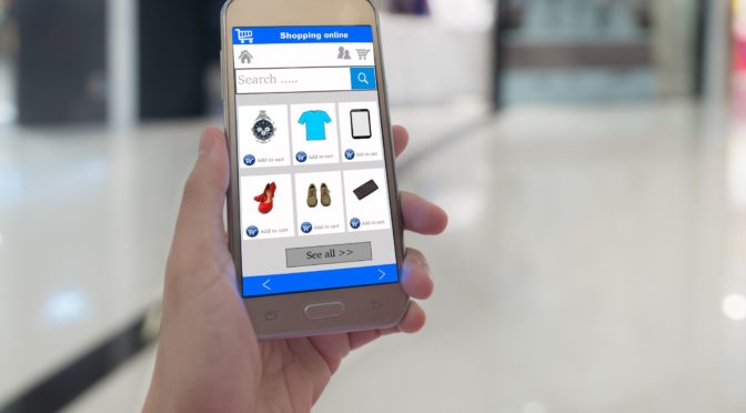 A hand holds a cell phone whose screen shows shopping app with a shopping cart symbol and various clothing items to choose.