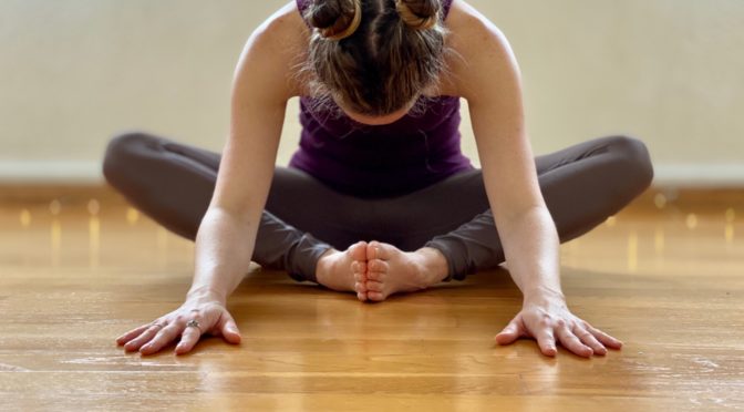 A woman in a yoga pose. She sits with her legs bent at the knee and her feet pressed together. She learns forward with her hands on the floor.