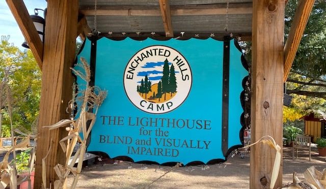 The Enchanted Hills Camp​ sign. The sign has a cyan blue background. At the top, inside of a white circle, it reads "Enchanted Hills Camp" with drawings of trees and clouds. Outside of the circle, at the bottom of the sign, it reads "The LightHouse for the Blind and Visually Impaired".