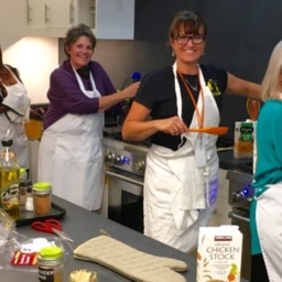 Students and instructors, all wearing white aprons, pose with cooking utensils in the LightHouse training kitchen