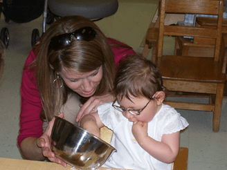 Pam sits behind a child wearing glasses who is reaching into a silver bowl Pam holds