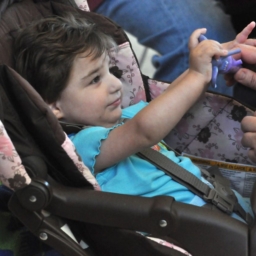 A baby sitting in a stroller seat plays with a tactile toy their parent is handing them