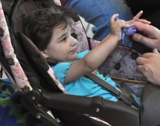 A baby sitting in a stroller seat plays with a tactile toy their parent is handing them