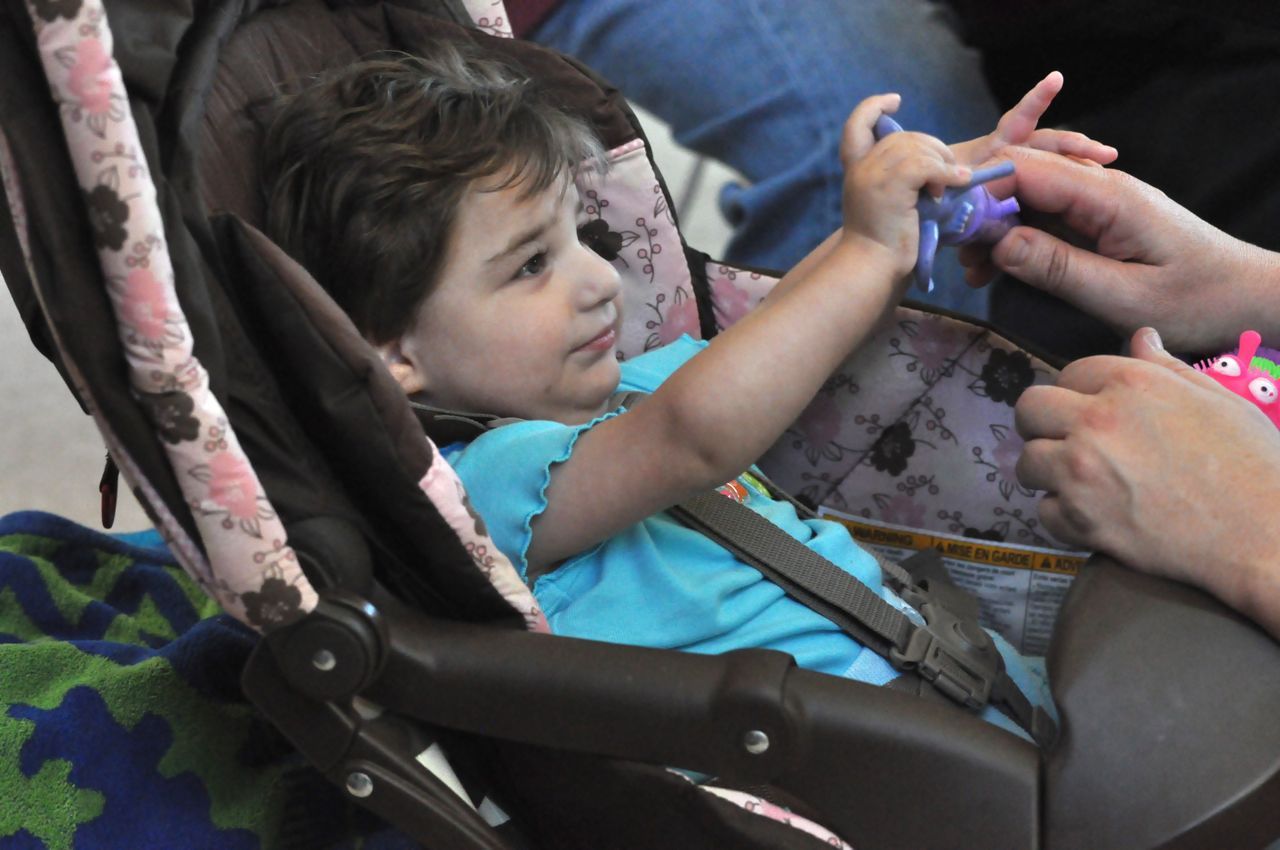 A baby sitting in a stroller seat plays with a tactile toy their parent is handing them