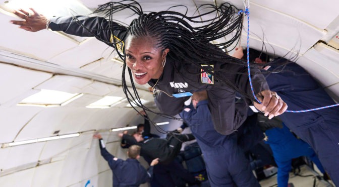 A black woman with long braids wearing a black one-piece flight suit floats suspended in mid-air inside a hollowed-out aircraft. She is loosely grasping a blue rope with her left hand