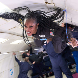 A black woman with long braids wearing a black one-piece flight suit floats suspended in mid-air inside a hollowed-out aircraft. She is loosely grasping a blue rope with her left hand