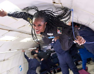 A black woman with long braids wearing a black one-piece flight suit floats suspended in mid-air inside a hollowed-out aircraft. She is loosely grasping a blue rope with her left hand