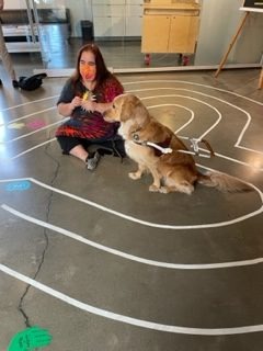 Maia Scott, wearing tie-dye, sits with her golden retriever guide dog in the middle of a heart shaped labyrinth she designed. She’s peeling a sun shaped sticker to place on the floor along with other colorful stickers created by the community at the Palo Alto Art Center.