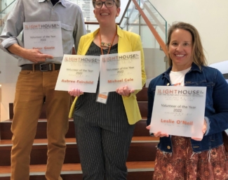 2022 Volunteers of the Year, Leslie O’Neil and Peter Goetz, stand on the LightHouse staircase with Allyson Ferrari, Volunteer Manager. Leslie and Peter are holding their Volunteer of the Year plaques. Note: Masks were temporarily removed to take this photo