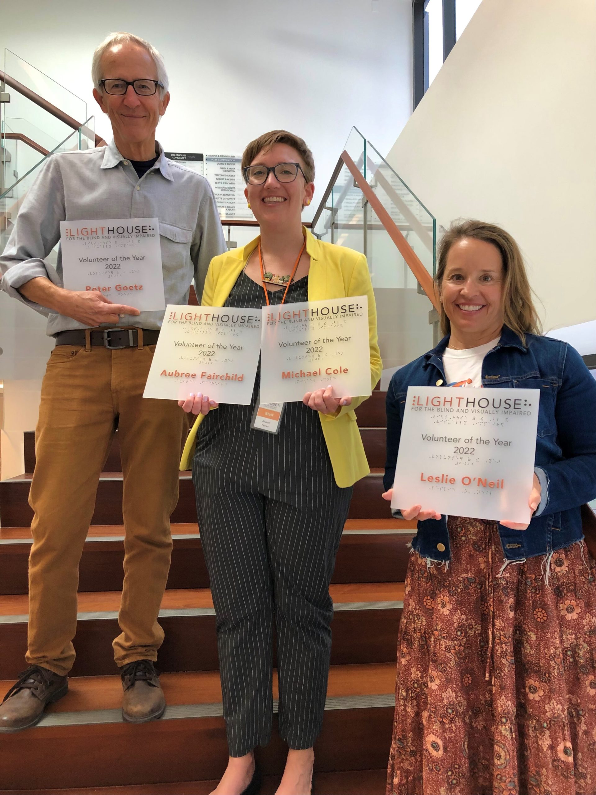 2022 Volunteers of the Year, Leslie O’Neil and Peter Goetz, stand on the LightHouse staircase with Allyson Ferrari, Volunteer Manager. Leslie and Peter are holding their Volunteer of the Year plaques. Note: Masks were temporarily removed to take this photo