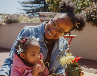 A mother cuddles with her daughter who is playing with a rainbow slinky toy. The mother is smiling and holding onto a silk flower planted in a pot