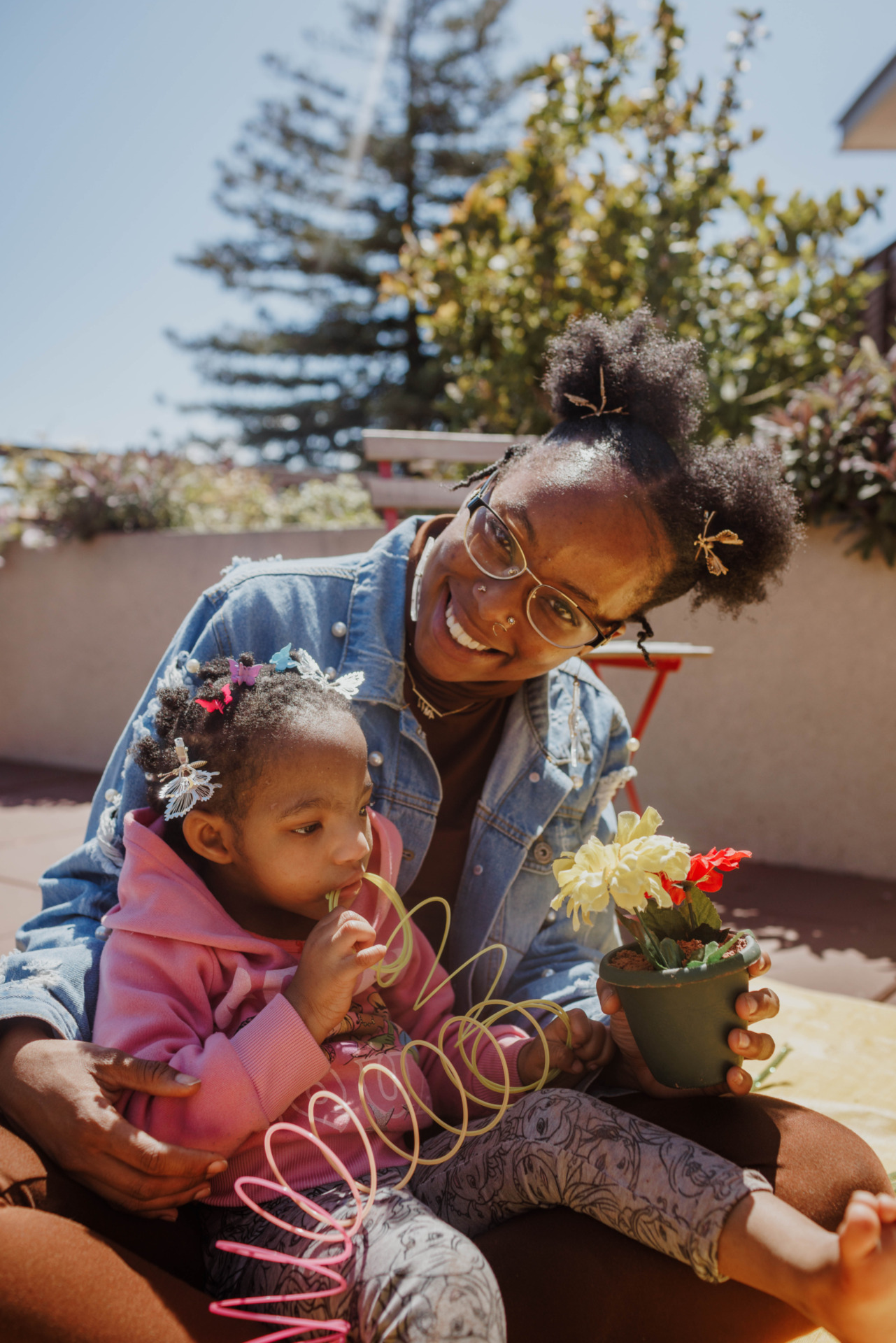 A mother cuddles with her daughter who is playing with a rainbow slinky toy. The mother is smiling and holding onto a silk flower planted in a pot