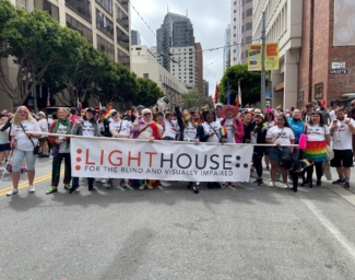 LightHouse for the Blind’s contingent in SF Pride 2022 making goofy faces at the camera. At least 15 people are holding the LightHouse banner across the road. All are wearing LightHouse’s Pride 2022 t-shirt. Participants are waving Pride flags, wearing a Pride flag as a cape, wearing a rainbow tutu, and a Guide Dog’s harness has a rainbow flag going across the bar. There are at least 30 people in the photo.