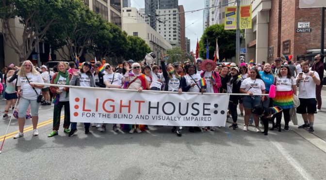 LightHouse for the Blind’s contingent in SF Pride 2022 making goofy faces at the camera. At least 15 people are holding the LightHouse banner across the road. All are wearing LightHouse’s Pride 2022 t-shirt. Participants are waving Pride flags, wearing a Pride flag as a cape, wearing a rainbow tutu, and a Guide Dog’s harness has a rainbow flag going across the bar. There are at least 30 people in the photo.