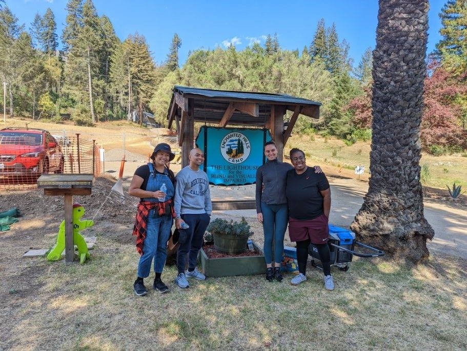 Four Verizon volunteers smile at the camera in front of the Enchanted Hills Camp sign.