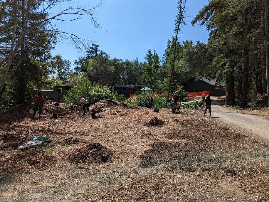 Five people with shovels, rakes, and wheelbarrows moving wood chips around. The back of the Hogan and two of the Lakeside Cabins are visible in the background.